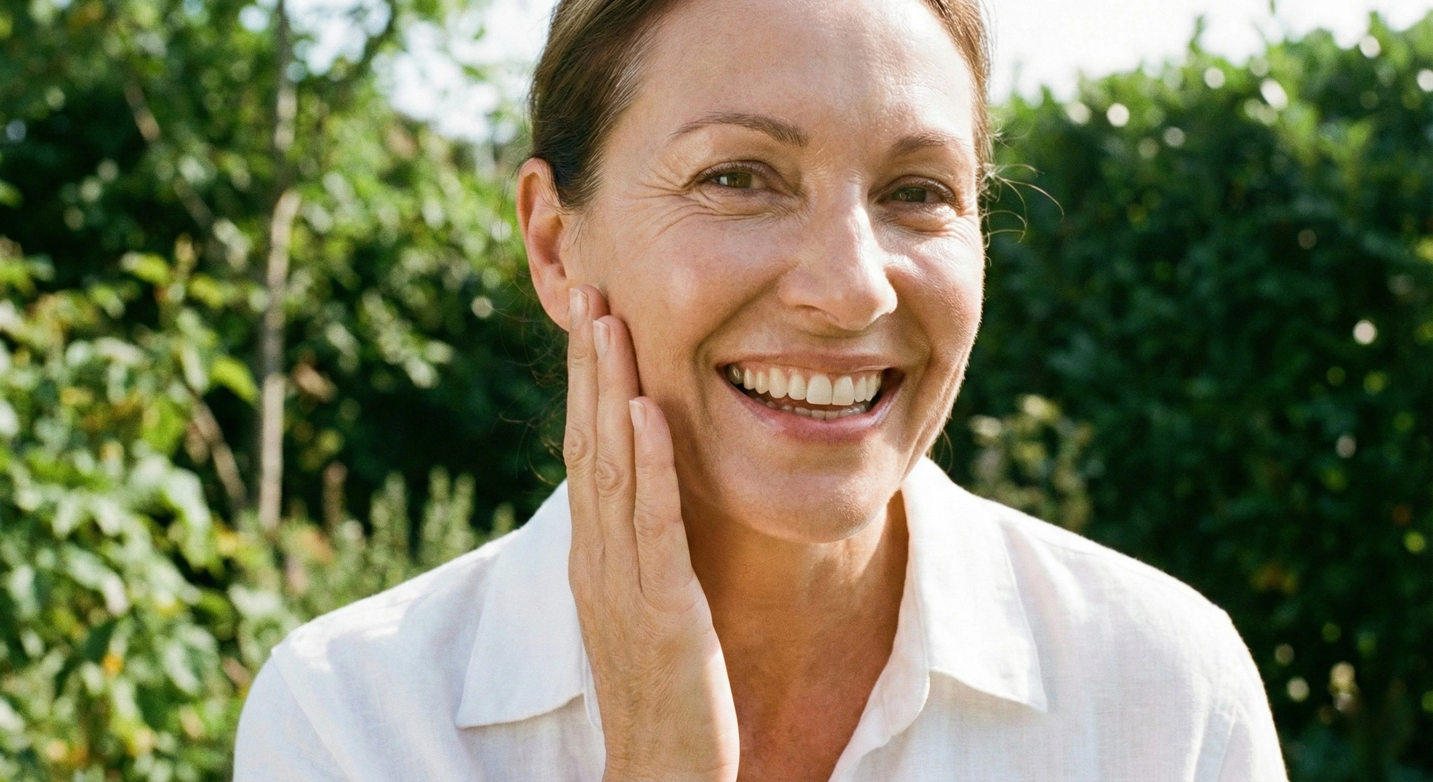 Donna di 50 anni sorridente con pelle luminosa e tonica che si tocca il viso con soddisfazione